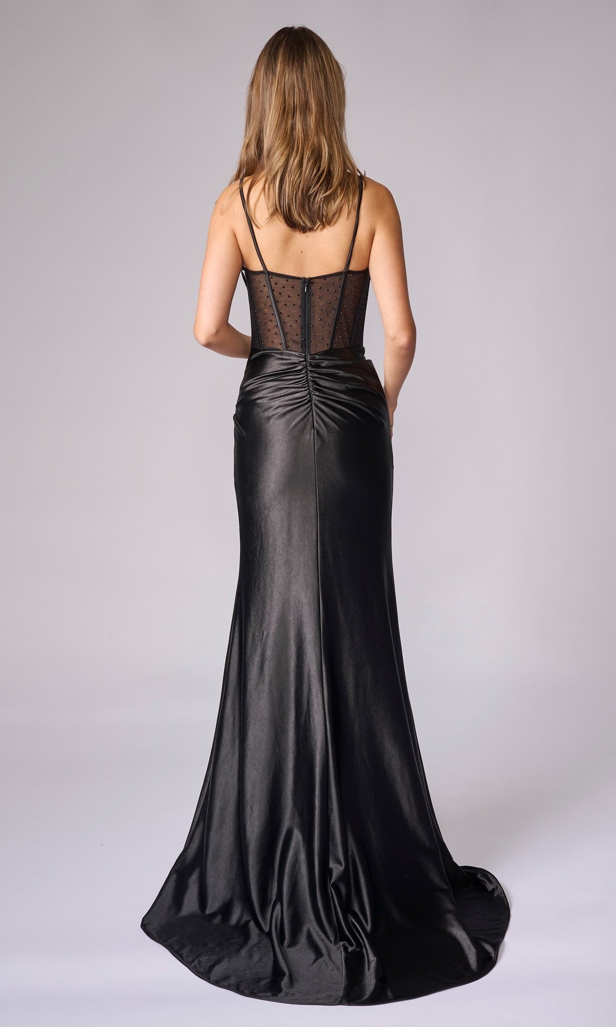 A woman faces away wearing the Nina Canacci 5410 long formal dress, a black satin gown with thin straps, sheer polka dot back, sheer-bodice effect, gathered seam, and a slight train for an elegant look.